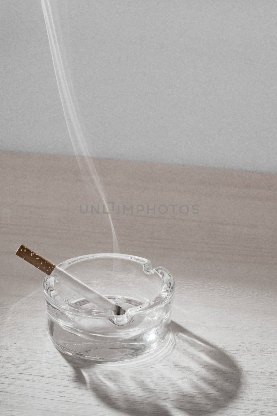 A reasently lid sigarette rests in a glass ashtray while the smoke floats up in the air.