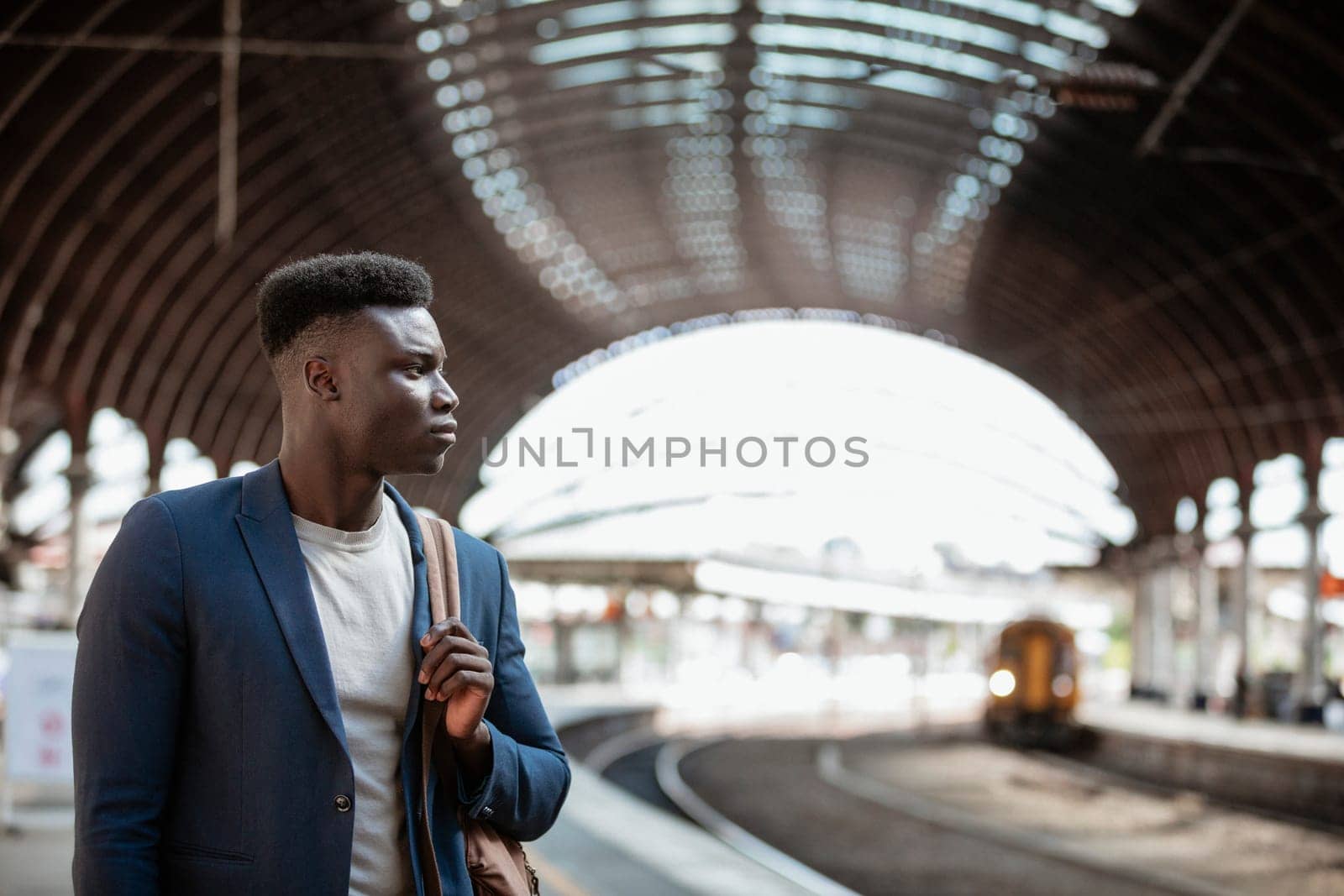 A businessman waiting for a train on the platform in a train station. He is holding a backpack over his shoulder.