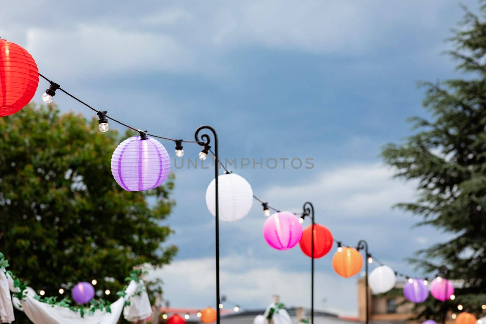 A wide shot of paper lanterns hanging up alongside a set of string lights at a garden party.