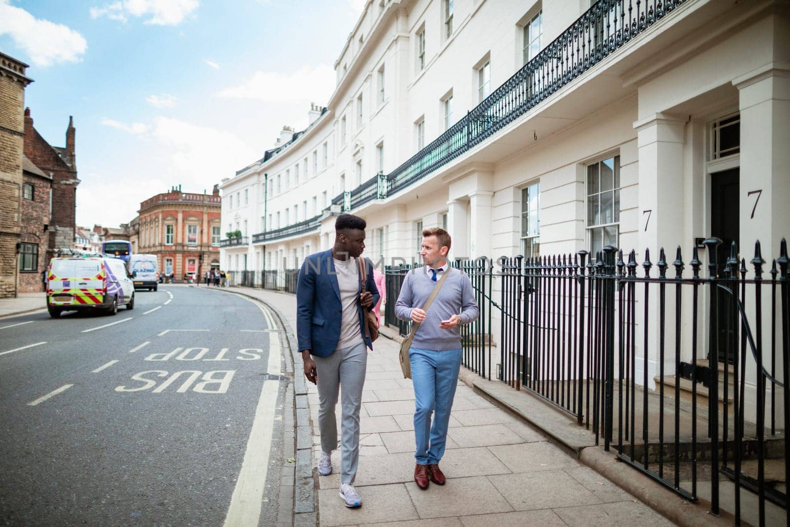 Two businessmen walking, on their way to work. They are talking and one man is holding a mobile phone ready to use it.