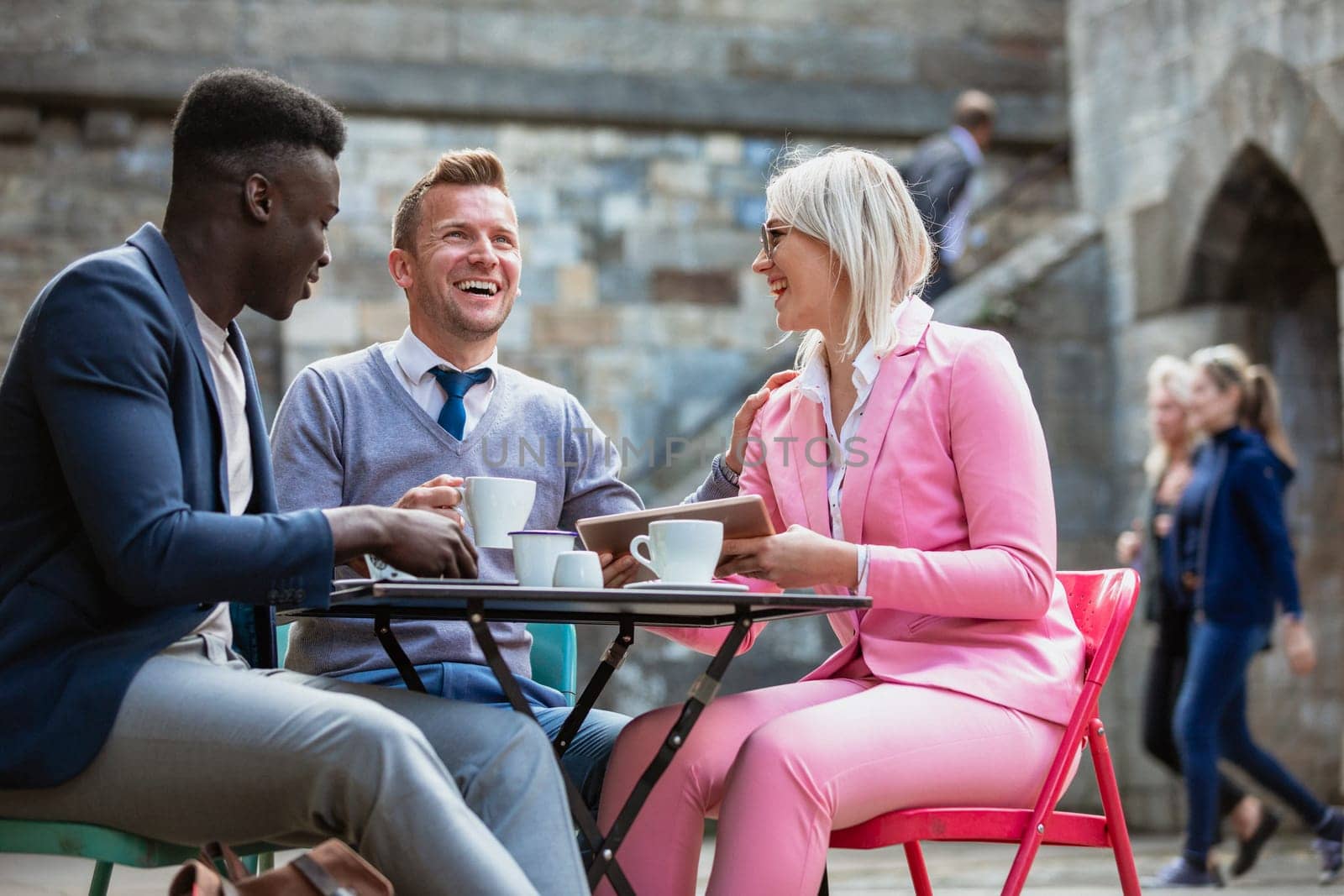 Two businessmen and a businesswoman sitting at an outdoor table in a city having a hot drink while laughing at something on a digital tablet. One man is touching the woman on the shoulder.
