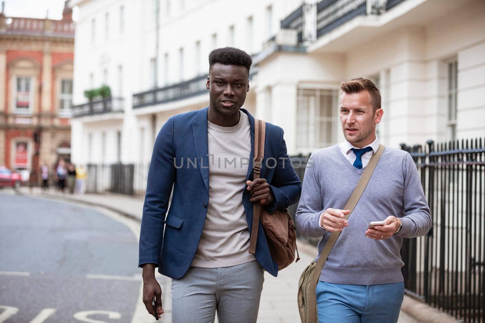 Two businessmen walking, on their way to work. They are talking and one man is holding a mobile phone ready to use it.