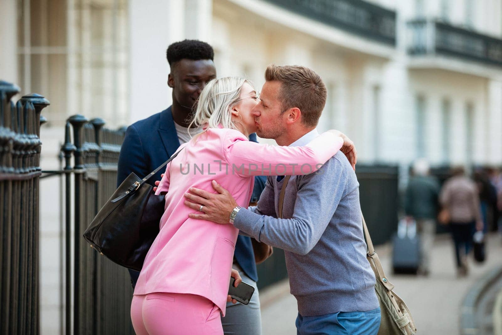 Two male and one female co-workers meeting up before work and greeting each other with a kiss on the cheek and hug.