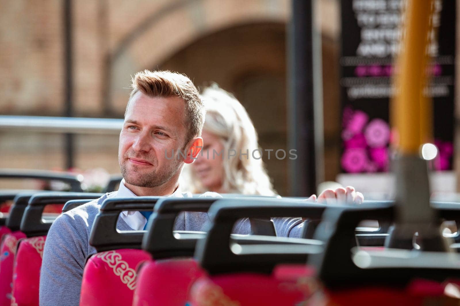 A businessman sitting by himself on an open-air bus  admiring the view. There is an unrecognisable woman behind him.