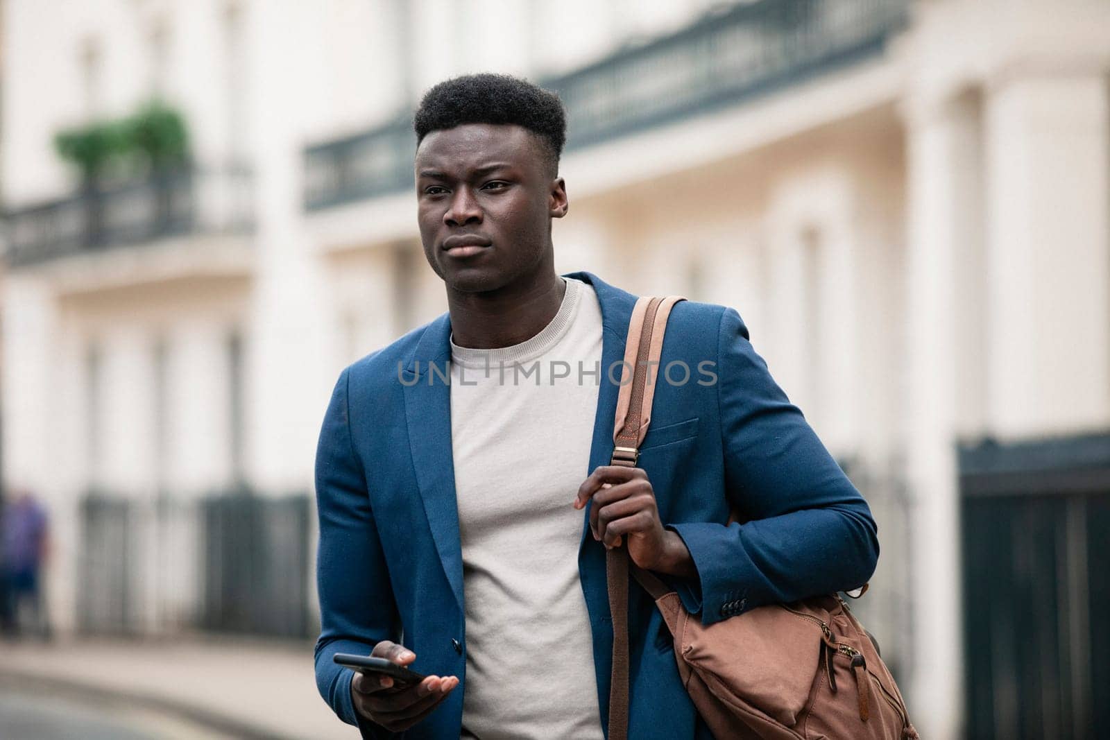 Businessman walking down a street on his way to work with a bag strap over his shoulder. He is holding his phone in one hand as if ready to browse the Internet but he is looking ahead.