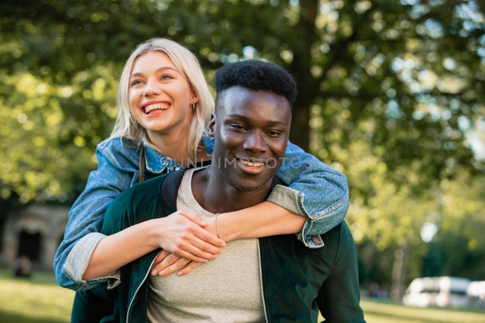 A close-up shot of a man giving a woman a piggyback lift through a park. They are both smiling.