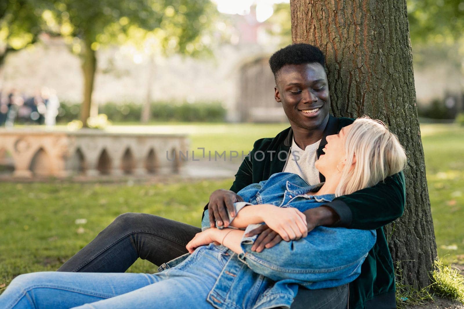 A man and woman sitting in a park against a tree. The man is holding her as she sits in between his legs. They are looking at each other while laughing.