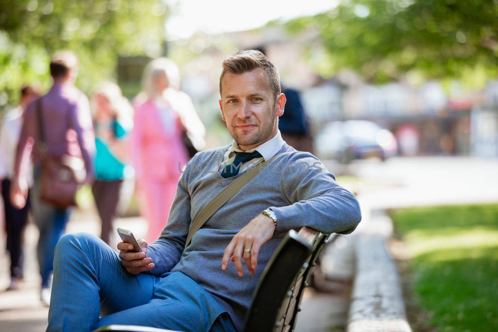A businessman sitting on a park bench while holding his mobile phone but looking at the camera.