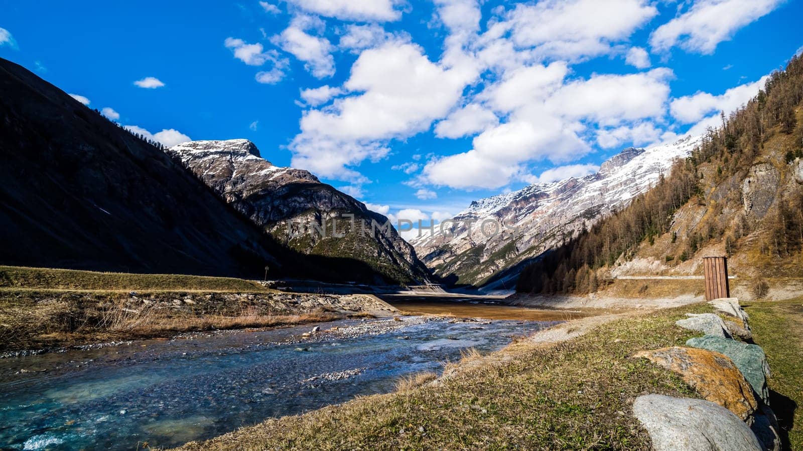 Beautiful mountain valley with stream, trees, small hut and dried Livigno lake, puffy clouds and blue sky in background, Alps, Italy