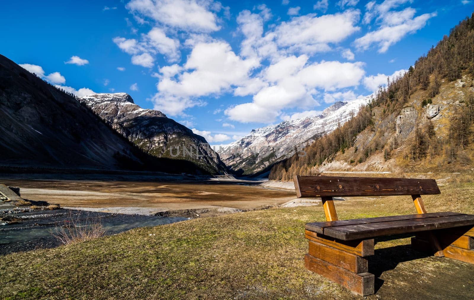 Beautiful mountain valley with stream, trees, wooden bench and dried Livigno lake, puffy clouds and blue sky in background, Alps, Italy