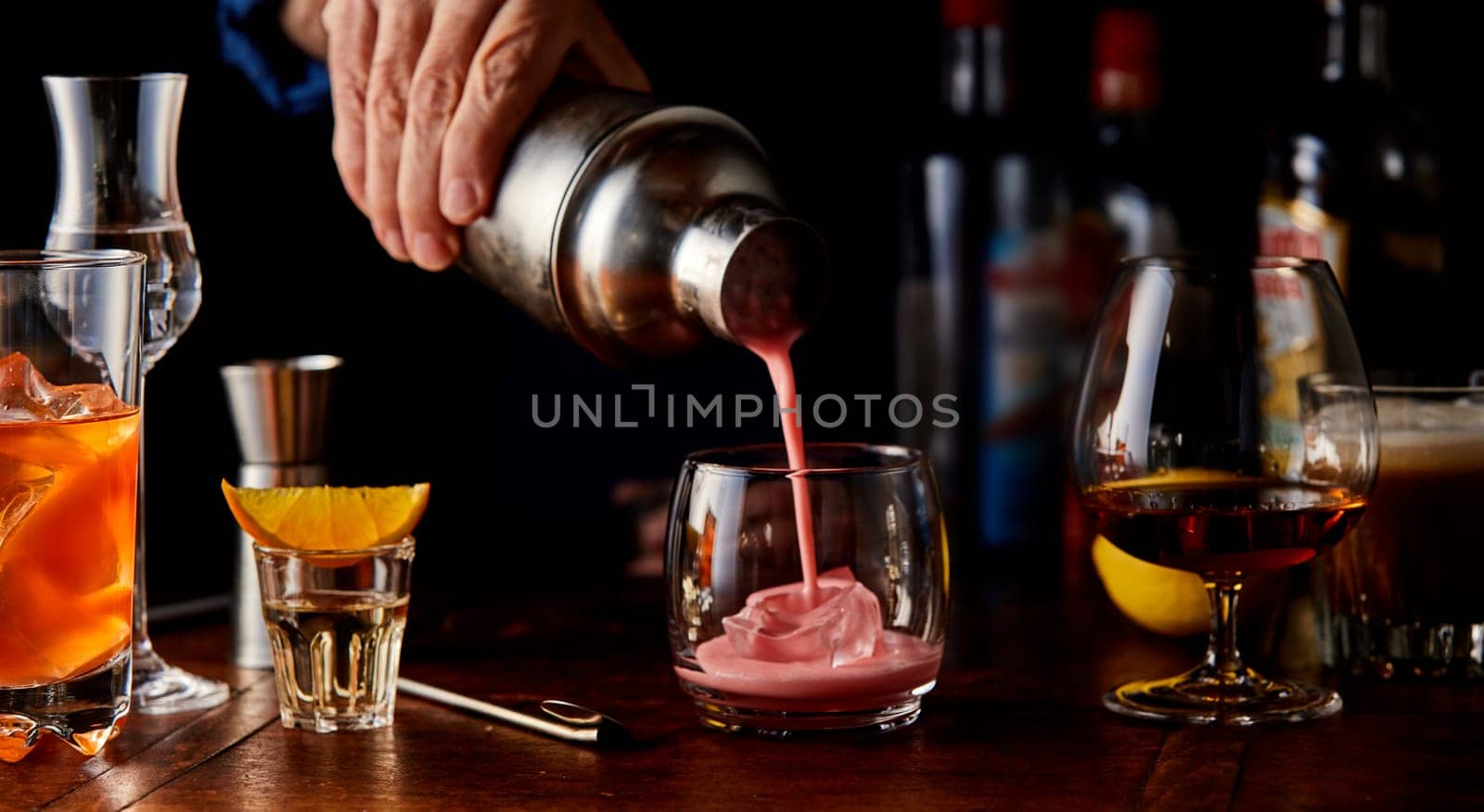Bartender pouring a fruity cocktail from a shaker on a wooden bar counter with an assortment of different alcoholic beverages in glasses