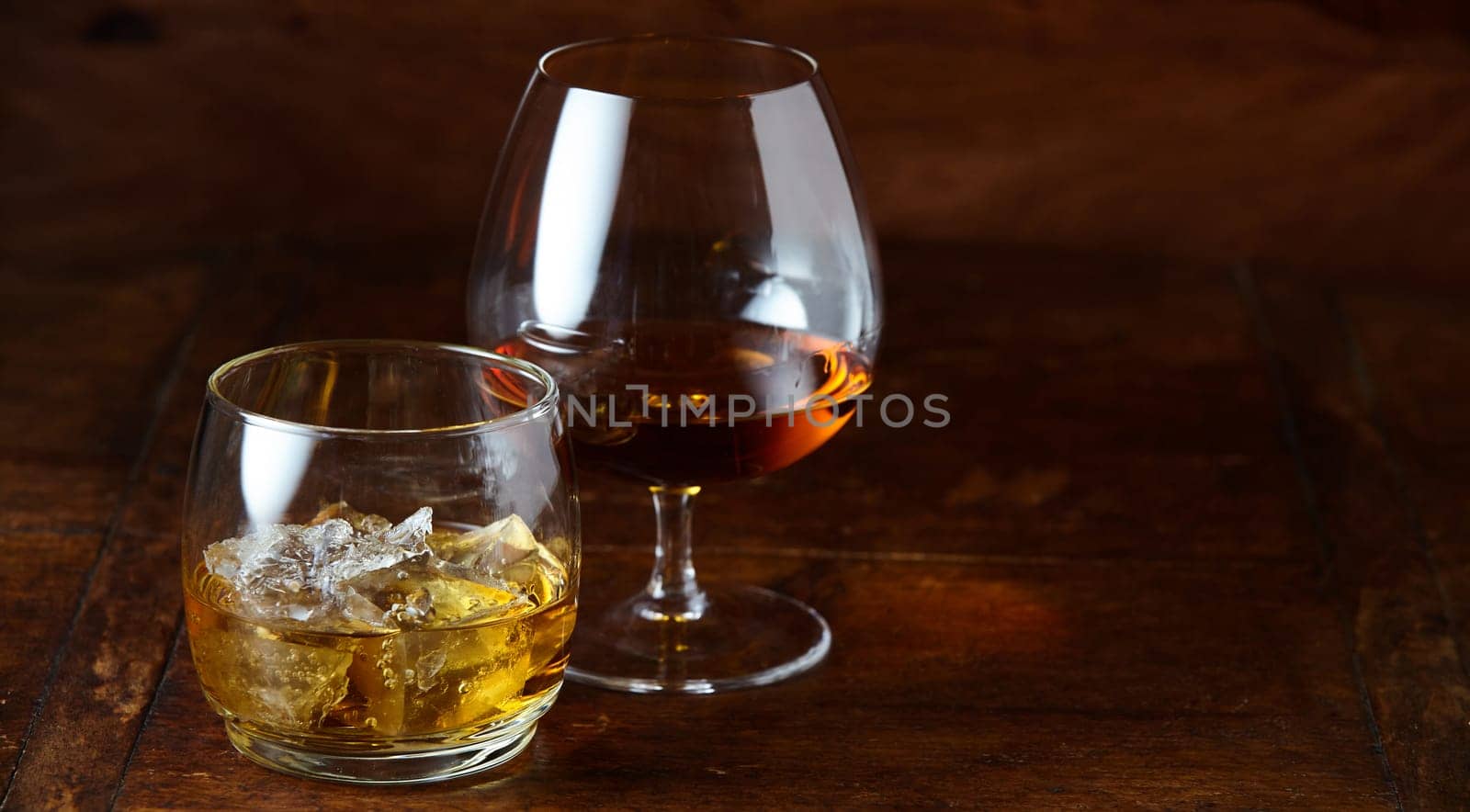 Whisky on the rocks and cognac in a snifter standing on a wooden bar counter at night with copy space