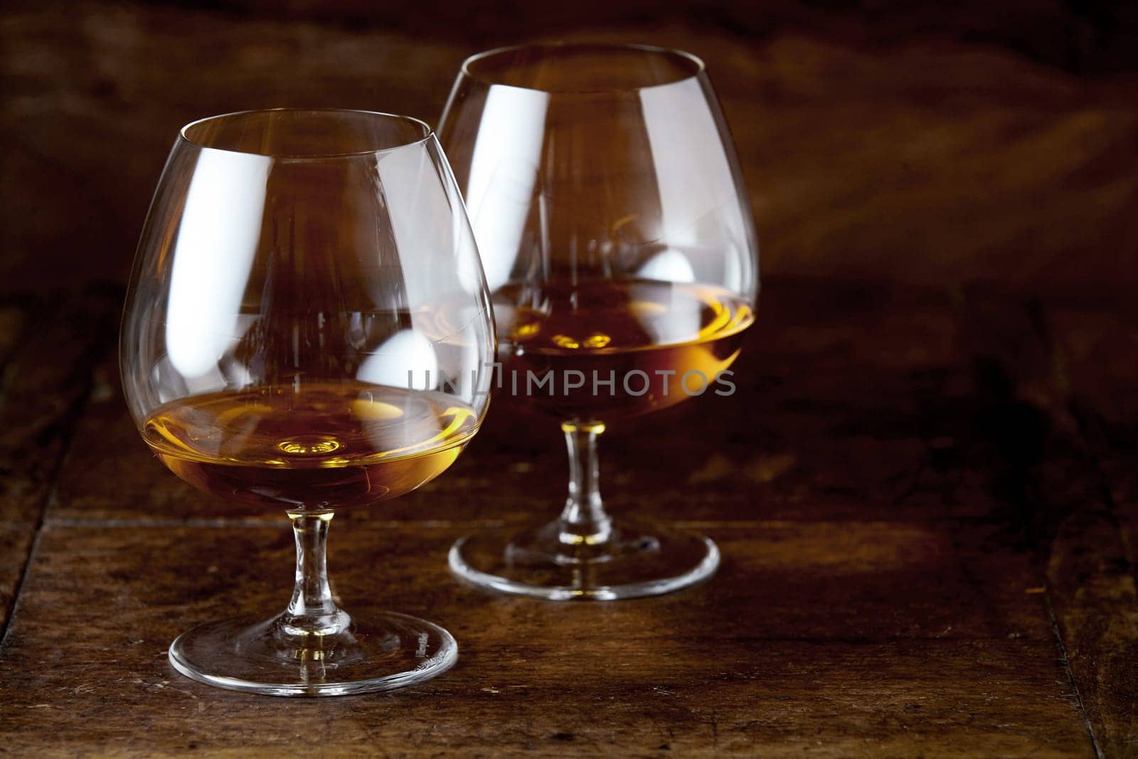 Two brandy snifter glasses with cognac in close-up on dark brown wooden counter surface with copy space