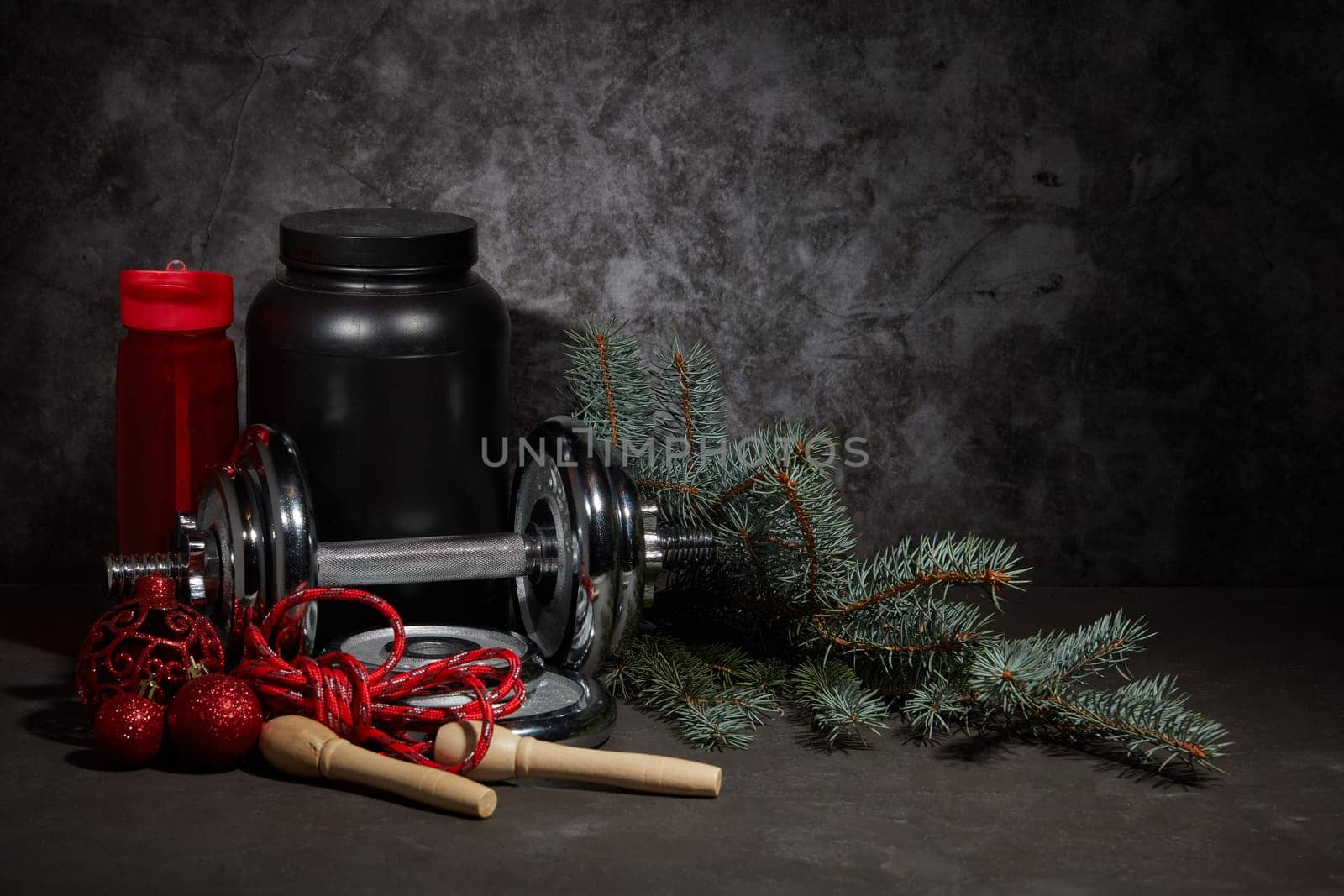Sports nutrition (supplements), dumbbell, bottle for water, jump rope and Christmas ornaments on dark grey background with copy space. Fitness, sport or healthy lifestyle concept. New Year. Christmas.