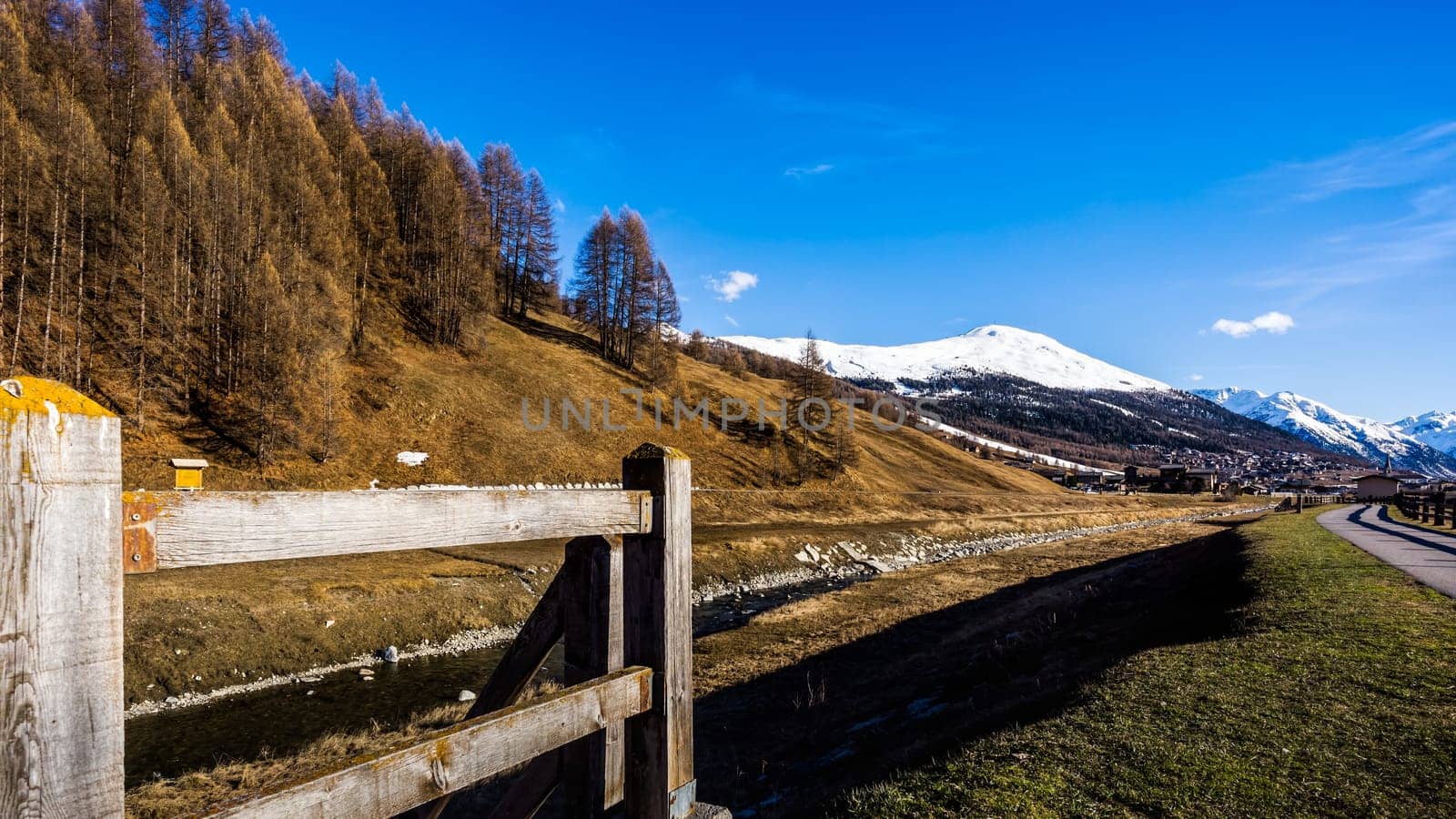 Beautiful mountain valley with stream, trees, wooden fence and footpath, Livingo village in background, Italy, Alps