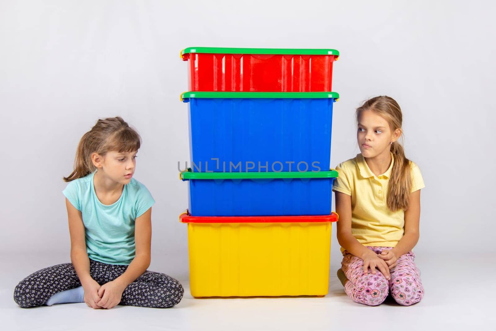 Two girls look at each other hiding behind large plastic boxes in the middle between them.