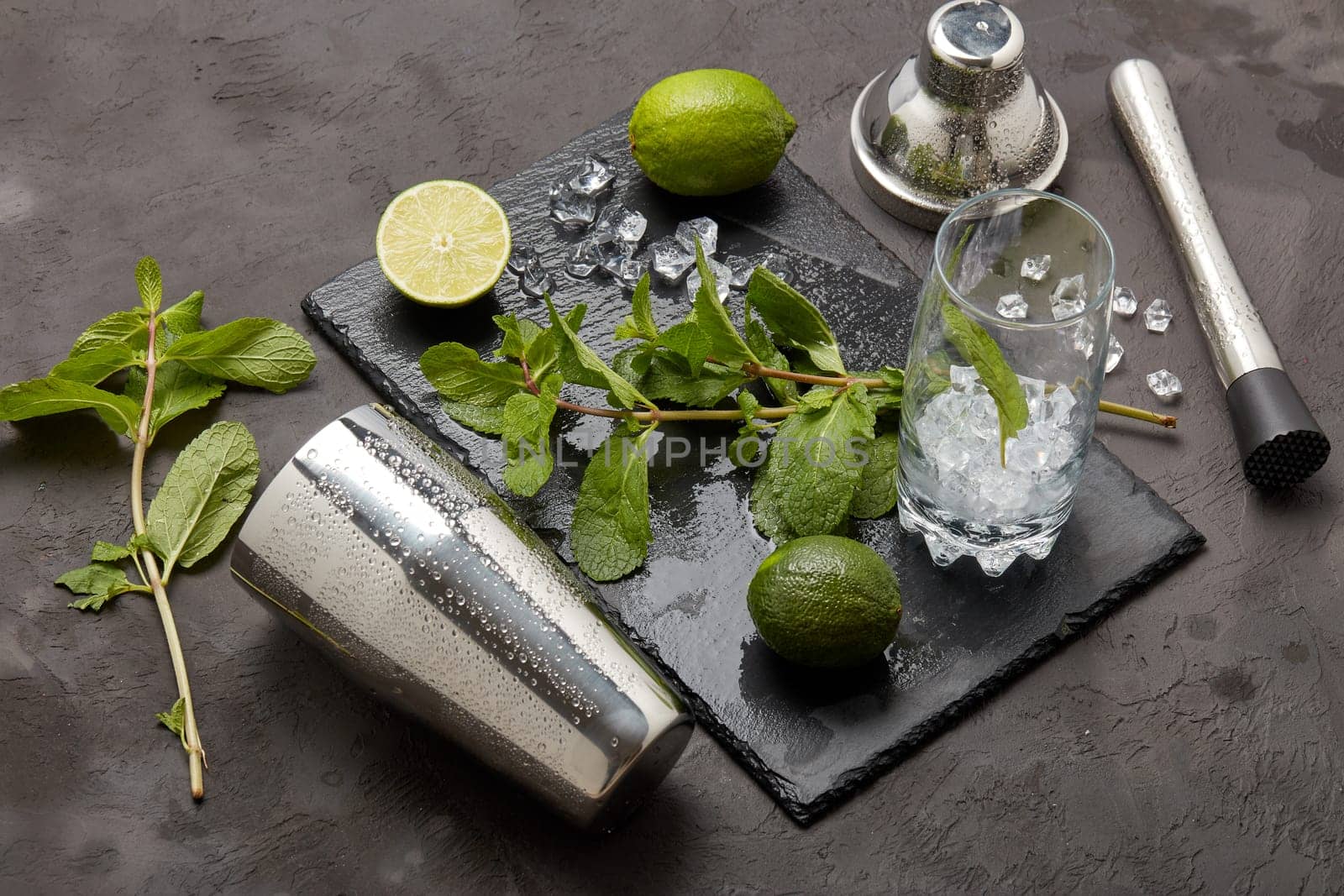 Set of bar tools: cocktail shaker, muddler and glass with ice, lime and mint on dark stone table. Top view. Bartender's tool. Mojito cocktail making.