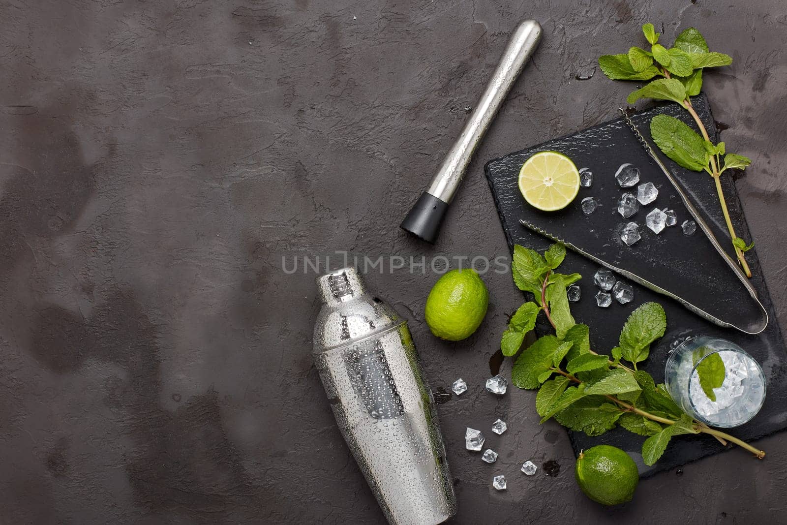 Set of bar tools: cocktail shaker, muddler, ice tongs and glass with ice, lime and mint on dark stone table. Top view with copy space. Bartender's tool. Mojito cocktail making.