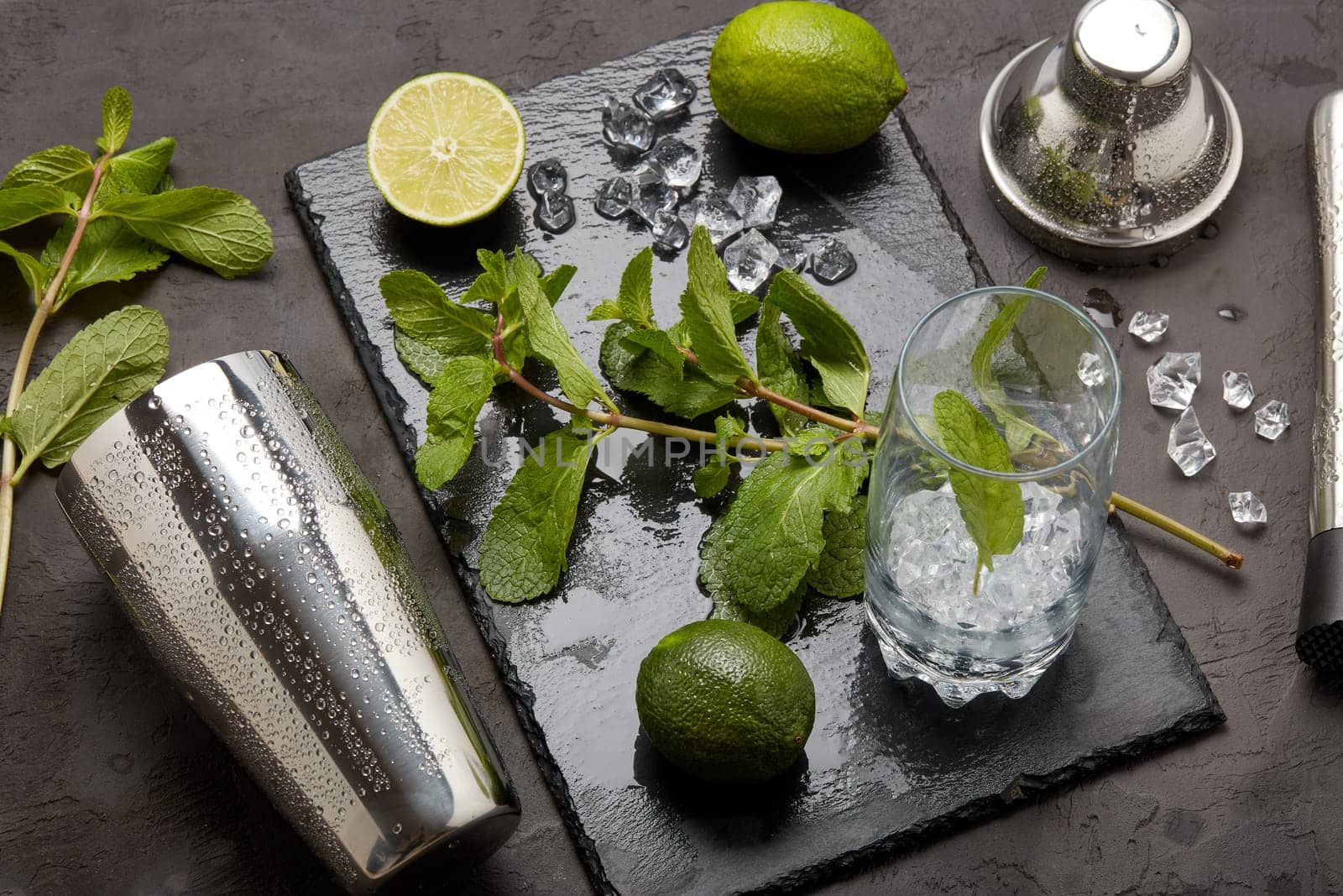 Set of bar tools: cocktail shaker, muddler and glass with ice, lime and mint on dark stone table. Top view. Bartender's tool. Mojito cocktail making.