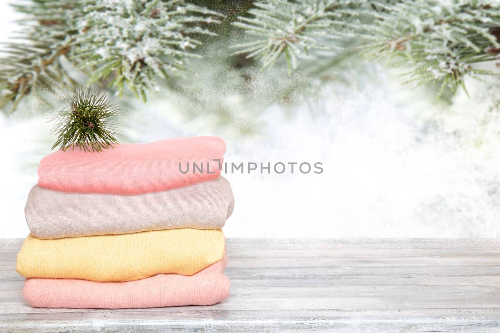 Pile of colorful knitted cozy warm cashmere sweaters on wooden table against blurred abstract snowy fir branches xmas background. Template for christmas sale.