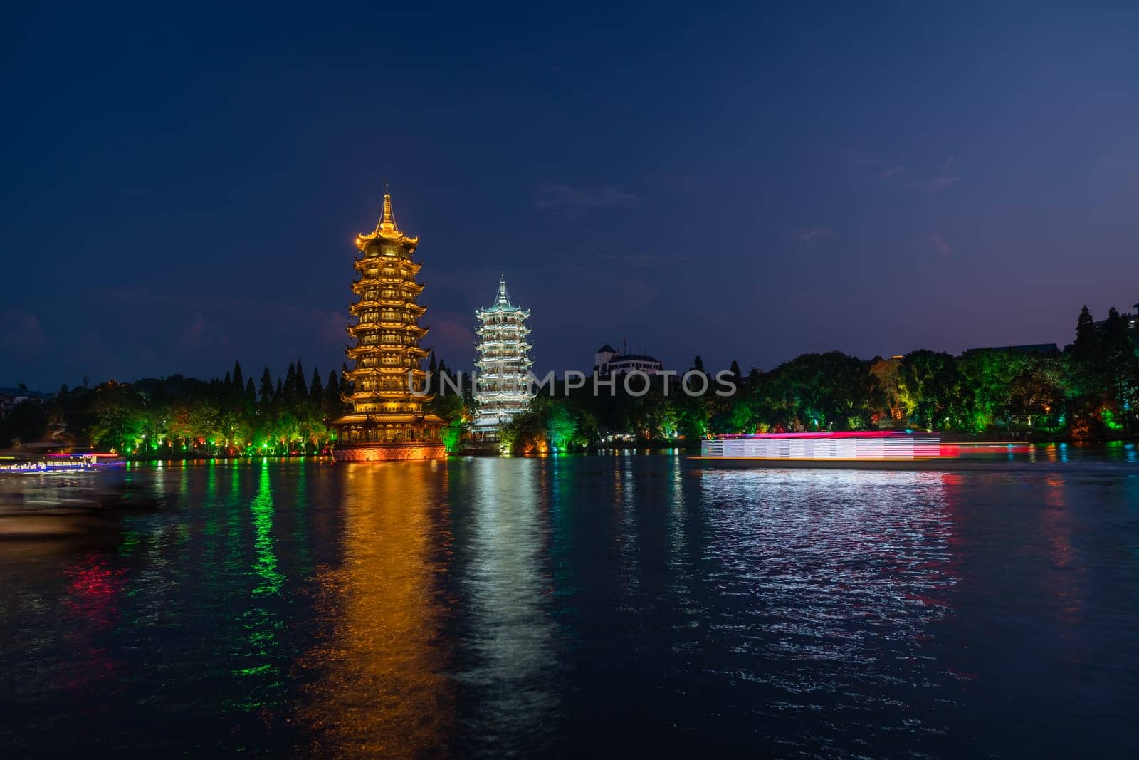 Tourist boats sailing in front of illuminated at night Sun and Moon Pagodas Towers at Shanhu or Shan Lake in Guilin town, Guangxi Province, China
