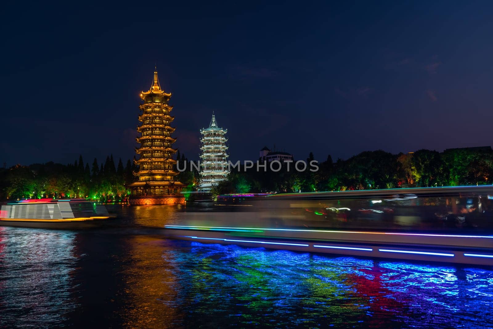 Tourist boats sailing in front of illuminated at night Sun and Moon Pagodas Towers at Shanhu or Shan Lake in Guilin town, Guangxi Province, China