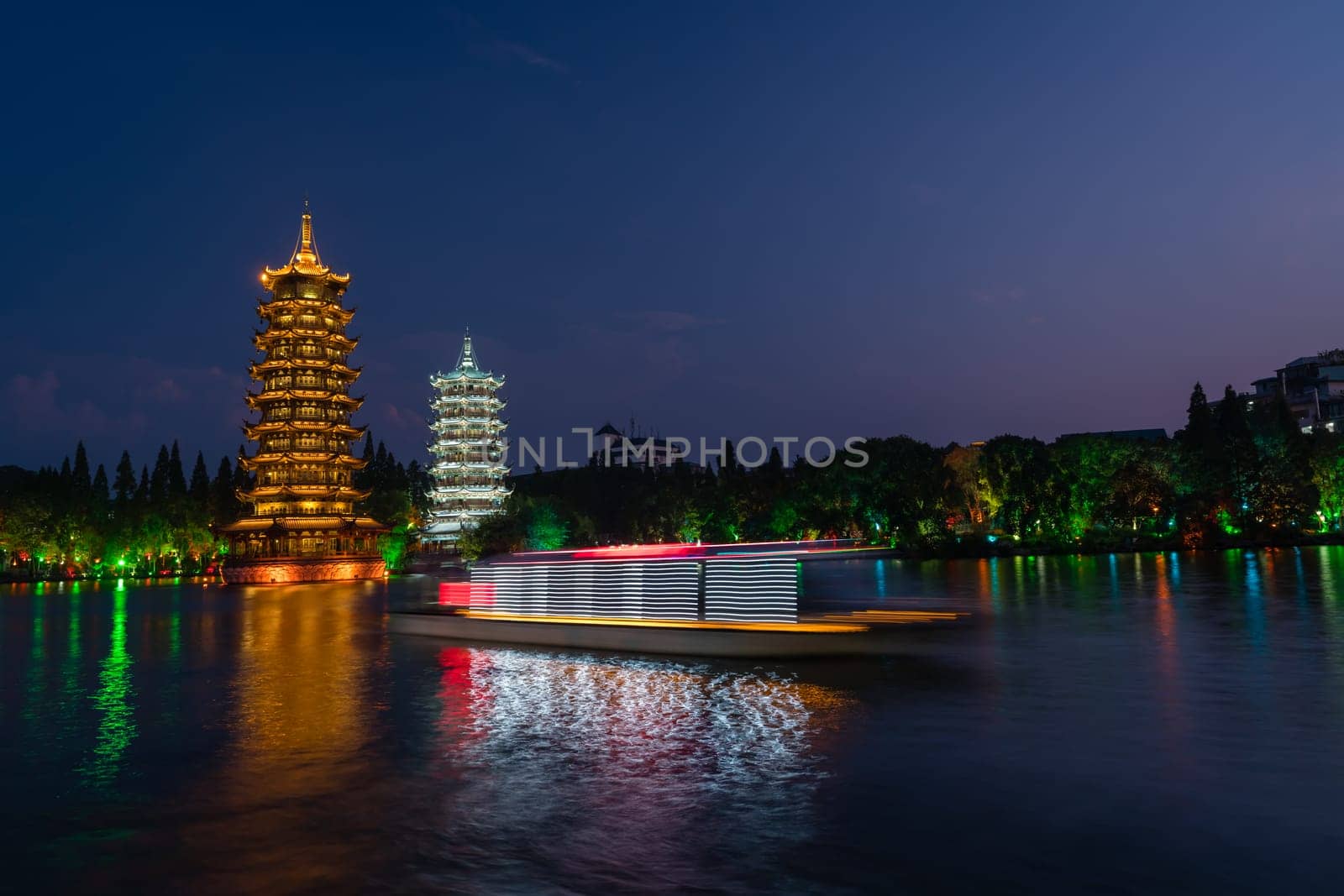 Tourist boats sailing in front of illuminated at night Sun and Moon Pagodas Towers at Shanhu or Shan Lake in Guilin town, Guangxi Province, China