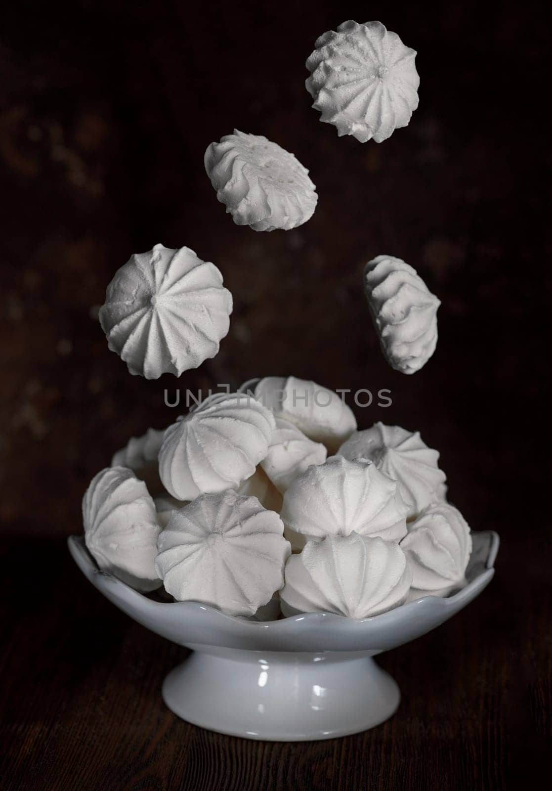 White meringues in a white porcelain vase on a dark brown background, with flying meringues on top, levitation