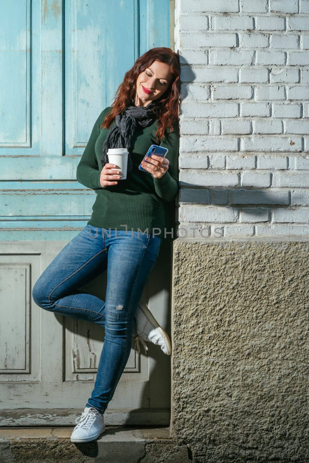 Young woman with red hair looking at her cellphone and drinking coffee from a takeout cup.