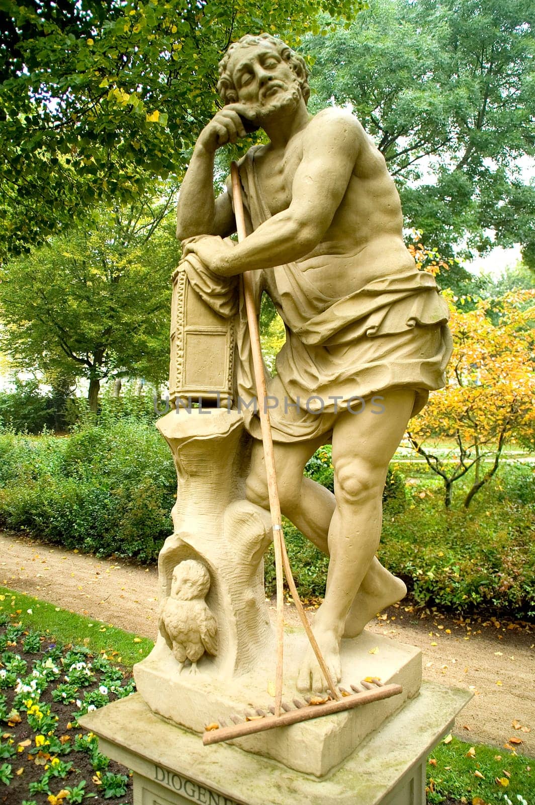 Diogenes sculpture in a relaxed position in a parc with a rake leaning unused in his arms.