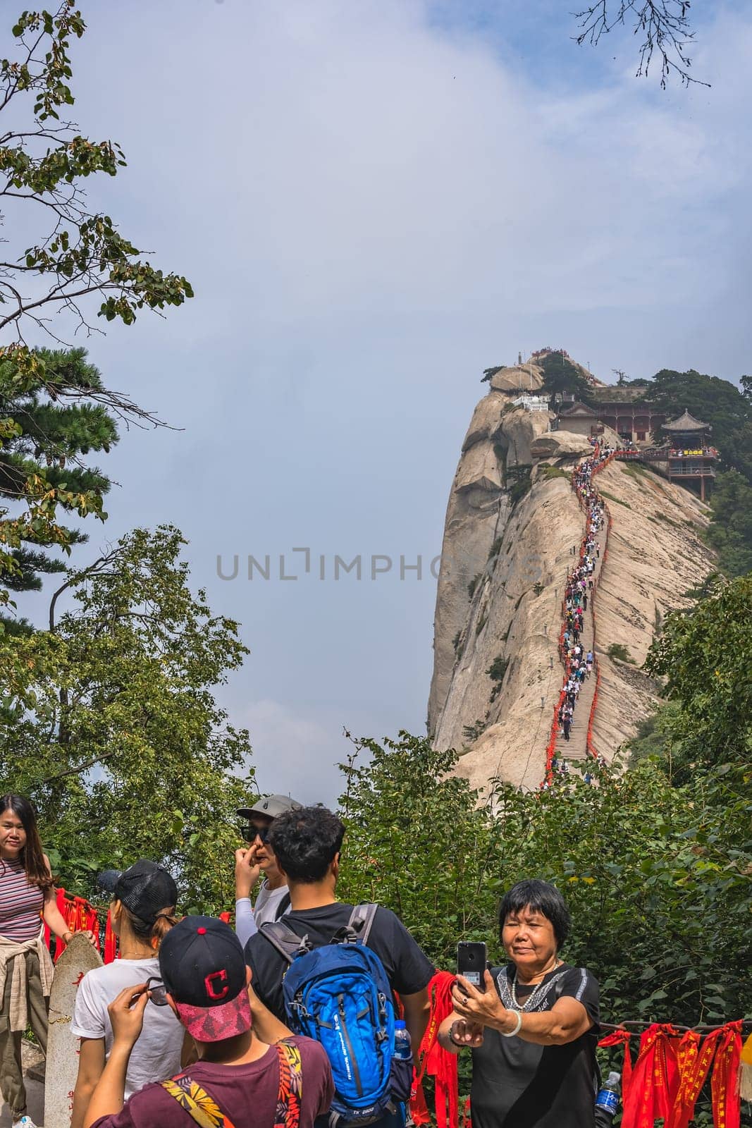 Huashan, China -  August 2019 : Chinese woman taking selfie picture on her phone with the crowds of tourists walking on a narrow ridge on a mountain trail to the West Peak in the background, Huashan mountain, Shaanxi Province