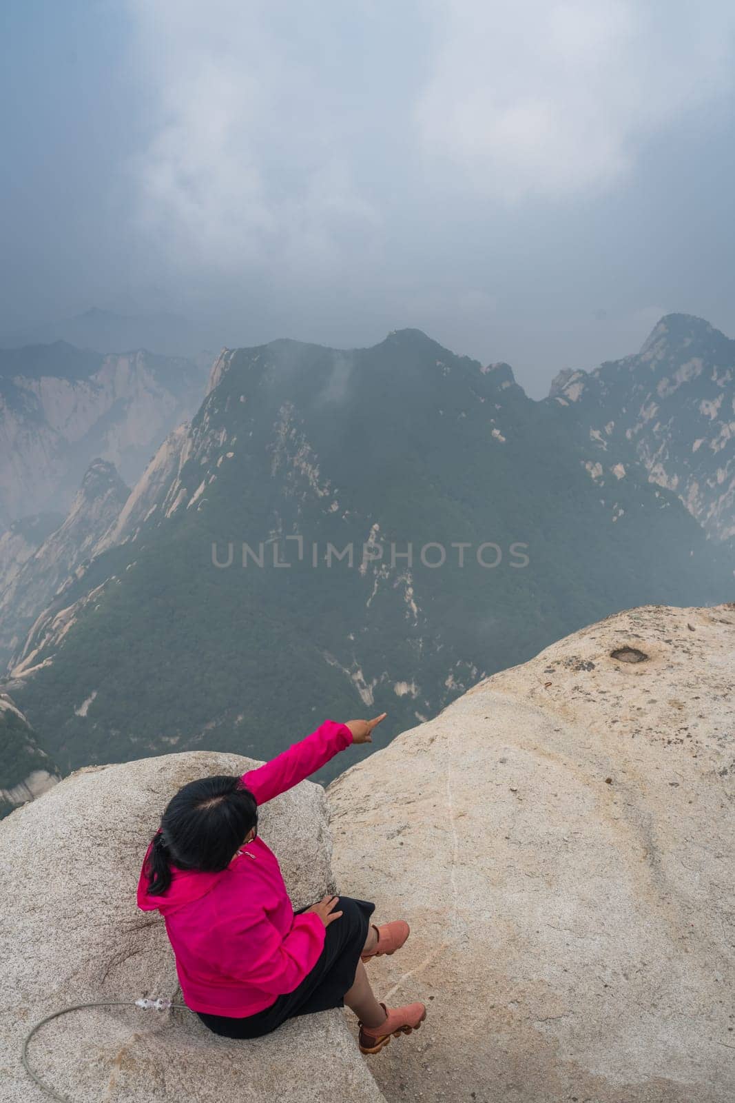 Huashan, China -  August 2019 : Tired chinese woman with sitting on a rock and pointing to the stunning landscape visible from the summit of the West Peak on Huashan mountain, Xian, Shaanxi Province