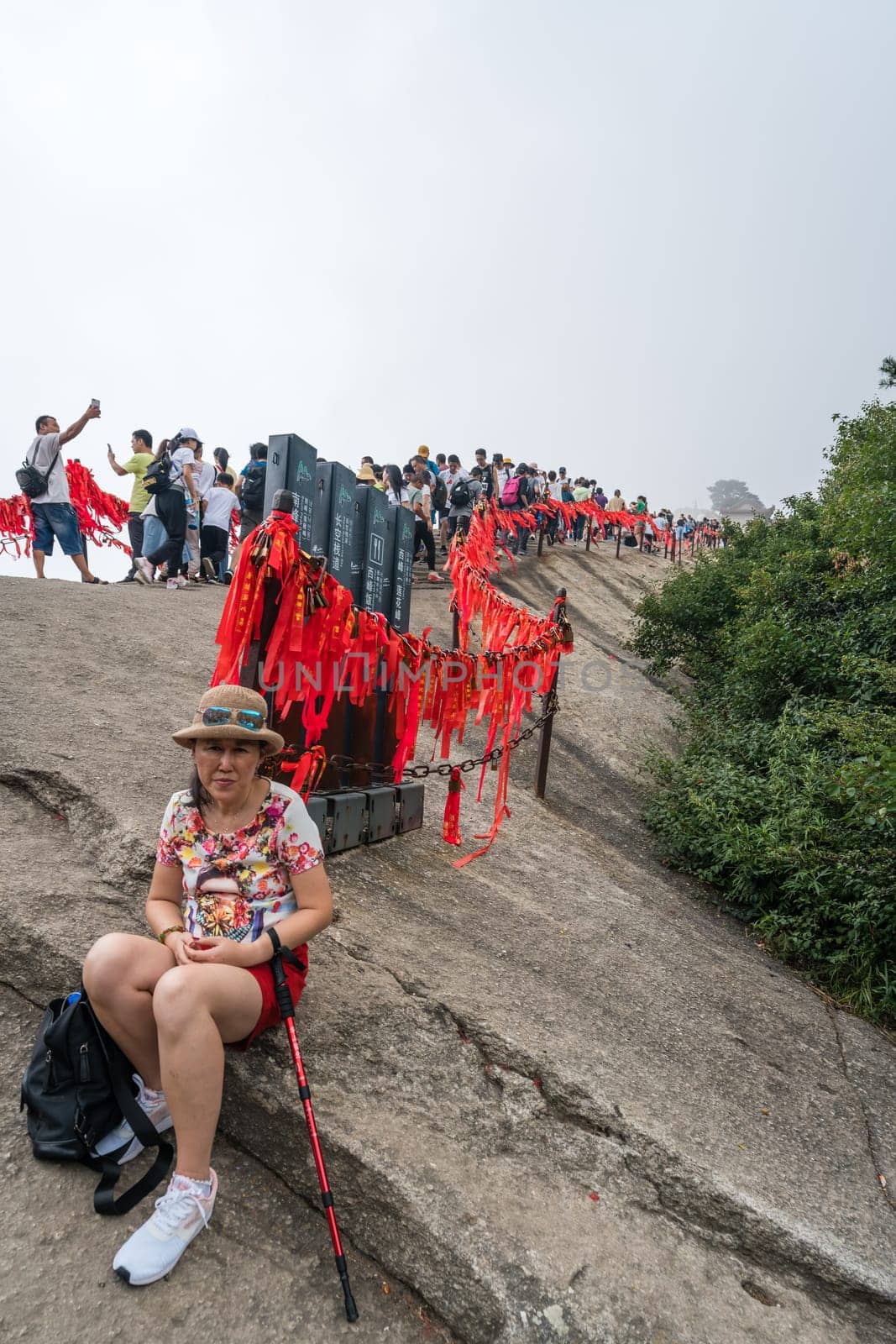 Huashan, China -  August 2019 : Tired chinese woman with a walking pole sitting on a rocky ground and resting on a mountain trail to the West Peak on Huashan mountain, Xian, Shaanxi Province