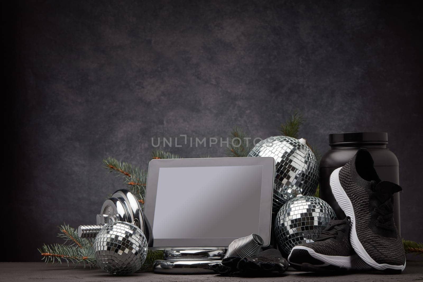 Sports nutrition, sports equipment, fir branch, Christmas ornament, blank computer tablet on dark grey background with copy space. Fitness, sport or healthy lifestyle concept. New Year and Christmas.