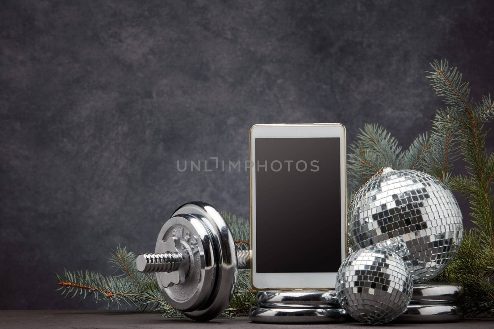 Sports equipment (dumbbell with weights), fir branch, Christmas ornament, blank computer tablet on dark grey background with copy space. Fitness, sport, healthy lifestyle concept. New Year. Christmas