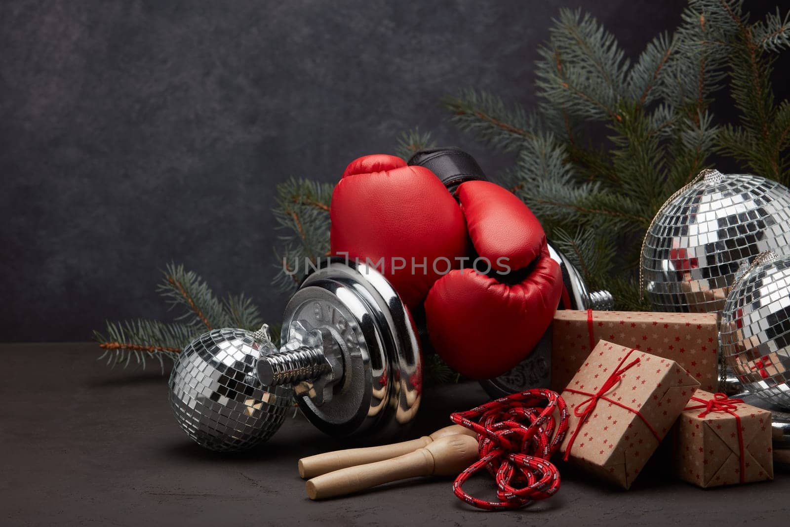 Sports equipment: dumbbell, boxing gloves, skipping rope, fir branch, Christmas ornament, gifts on dark grey background with copy space. Fitness, sport, healthy lifestyle concept. New Year. Christmas
