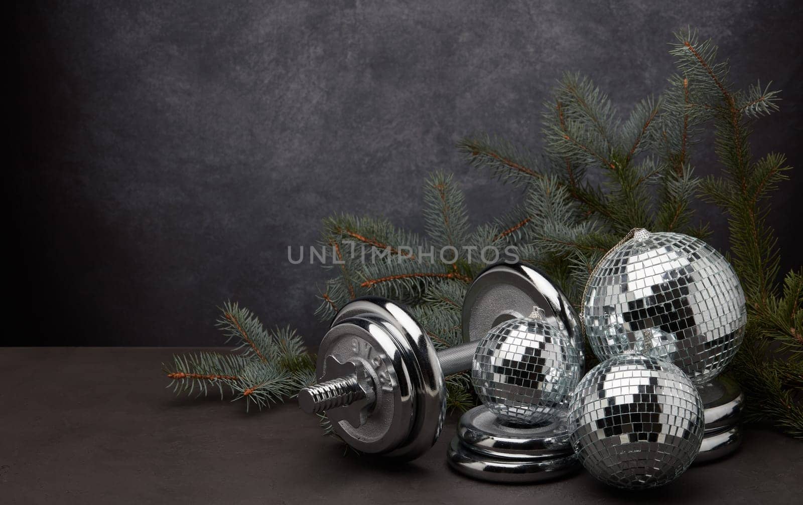 Sports equipment (dumbbell with weights), fir branch, Christmas ornament on dark grey background with copy space. Fitness, sport or healthy lifestyle concept. New Year and Christmas.