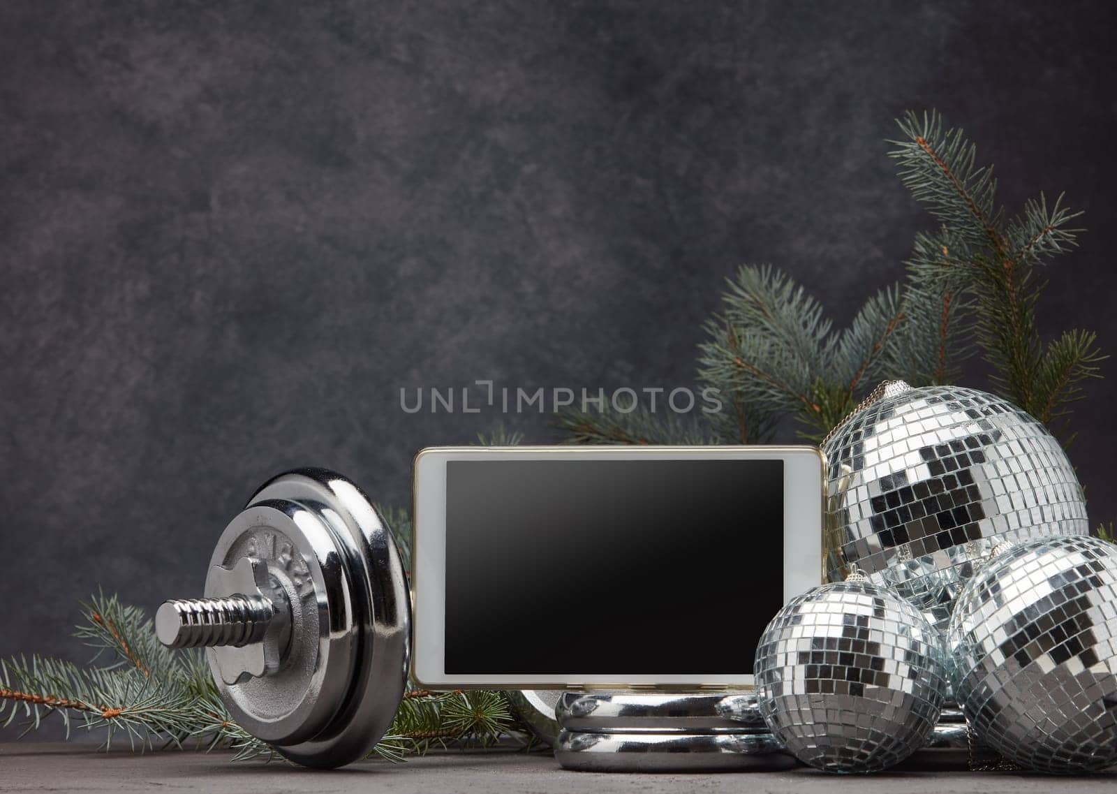 Sports equipment (dumbbell with weights), fir branch, Christmas ornament, blank computer tablet on dark grey background with copy space. Fitness, sport, healthy lifestyle concept. New Year. Christmas