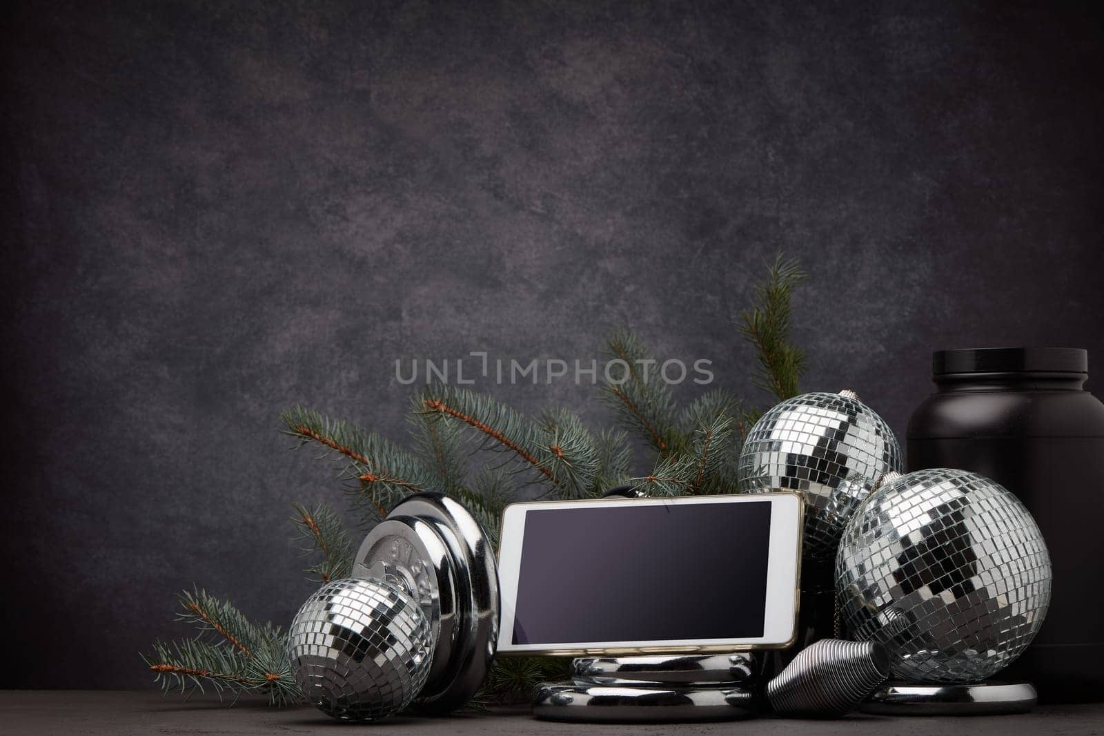 Sports nutrition, sports equipment, fir branch, Christmas ornament, blank computer tablet on dark grey background with copy space. Fitness, sport or healthy lifestyle concept. New Year and Christmas.