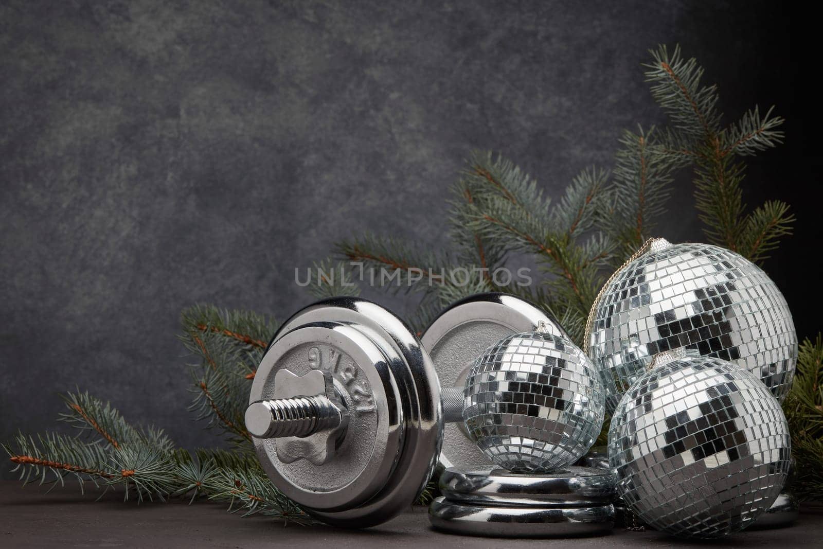 Sports equipment (dumbbell with weights), fir branch, Christmas ornament on dark grey background with copy space. Fitness, sport or healthy lifestyle concept. New Year and Christmas.