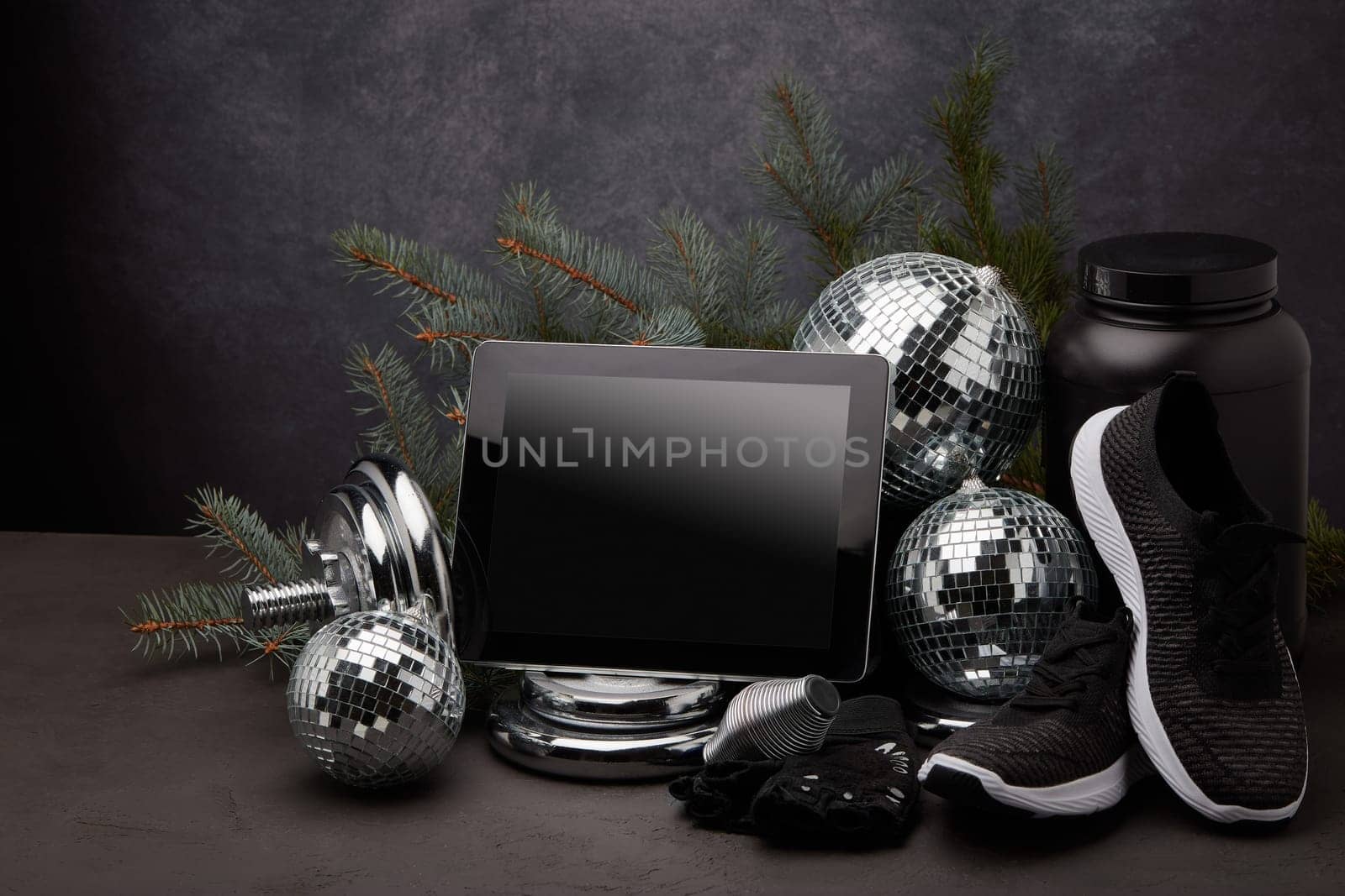 Sports nutrition, sports equipment, fir branch, Christmas ornament, blank computer tablet on dark grey background with copy space. Fitness, sport or healthy lifestyle concept. New Year and Christmas.