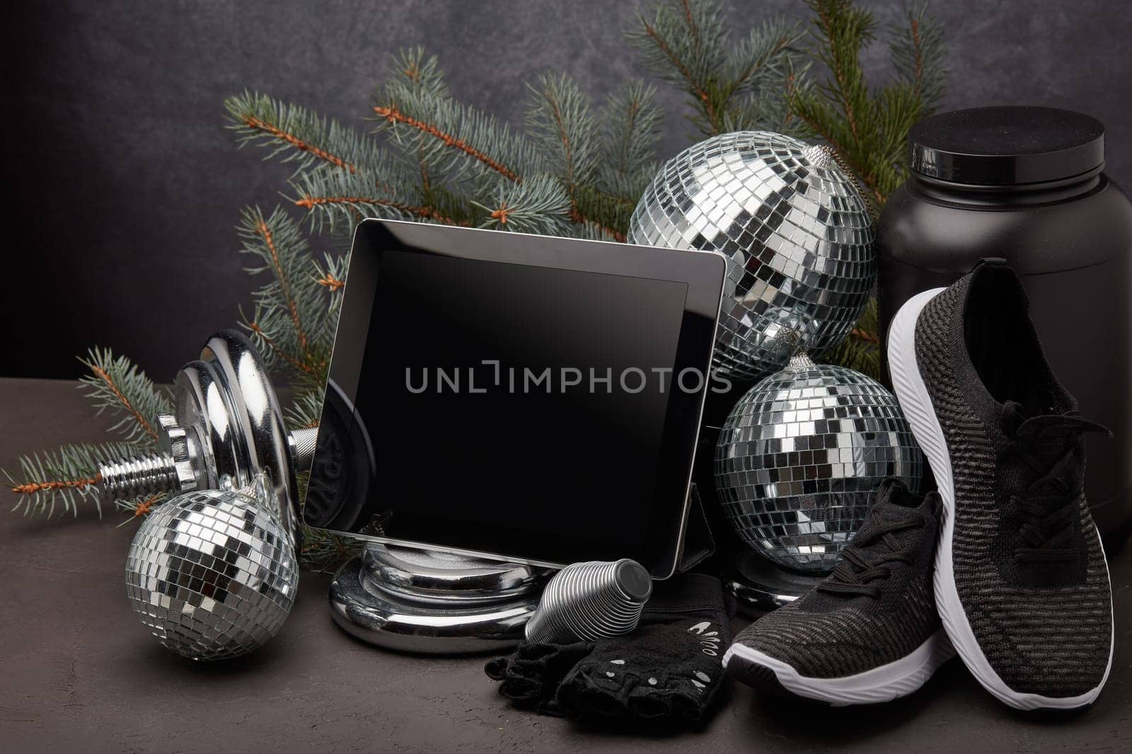 Sports nutrition, sports equipment, fir branch, Christmas ornament, blank computer tablet on dark grey background with copy space. Fitness, sport or healthy lifestyle concept. New Year and Christmas.