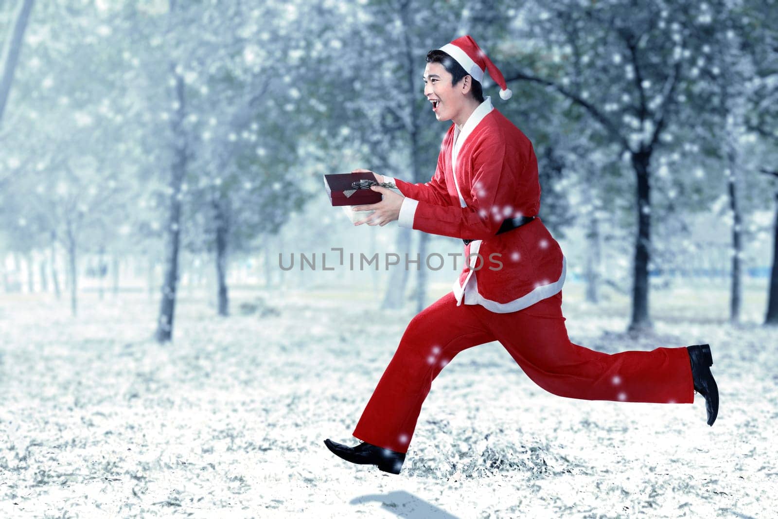 Asian man in Santa costume running while holding the gift box with snowy trees background
