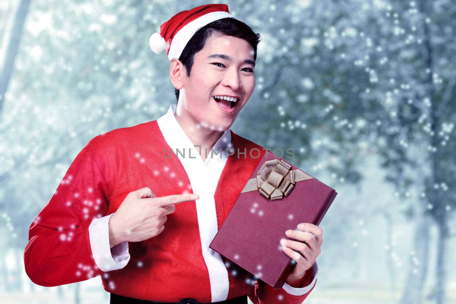 Asian man in Santa costume holding gift box with snowy trees background