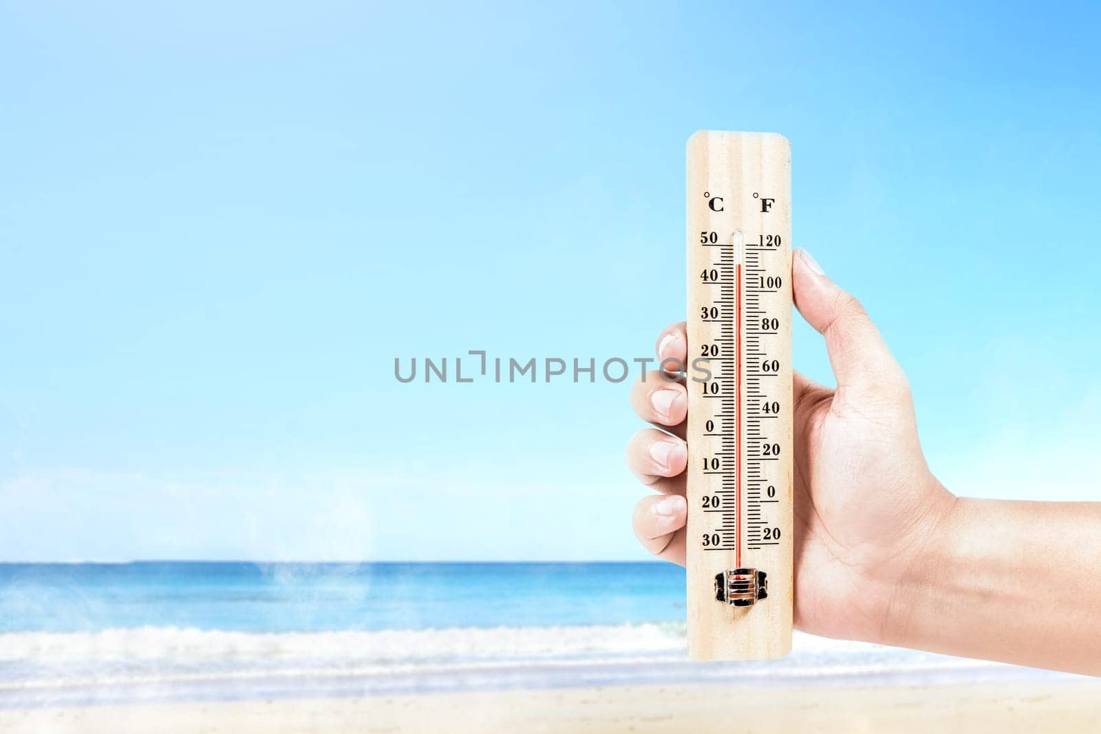 Hand holding thermometer measuring the temperature on the beach with a blue sky background. Heatwave concept