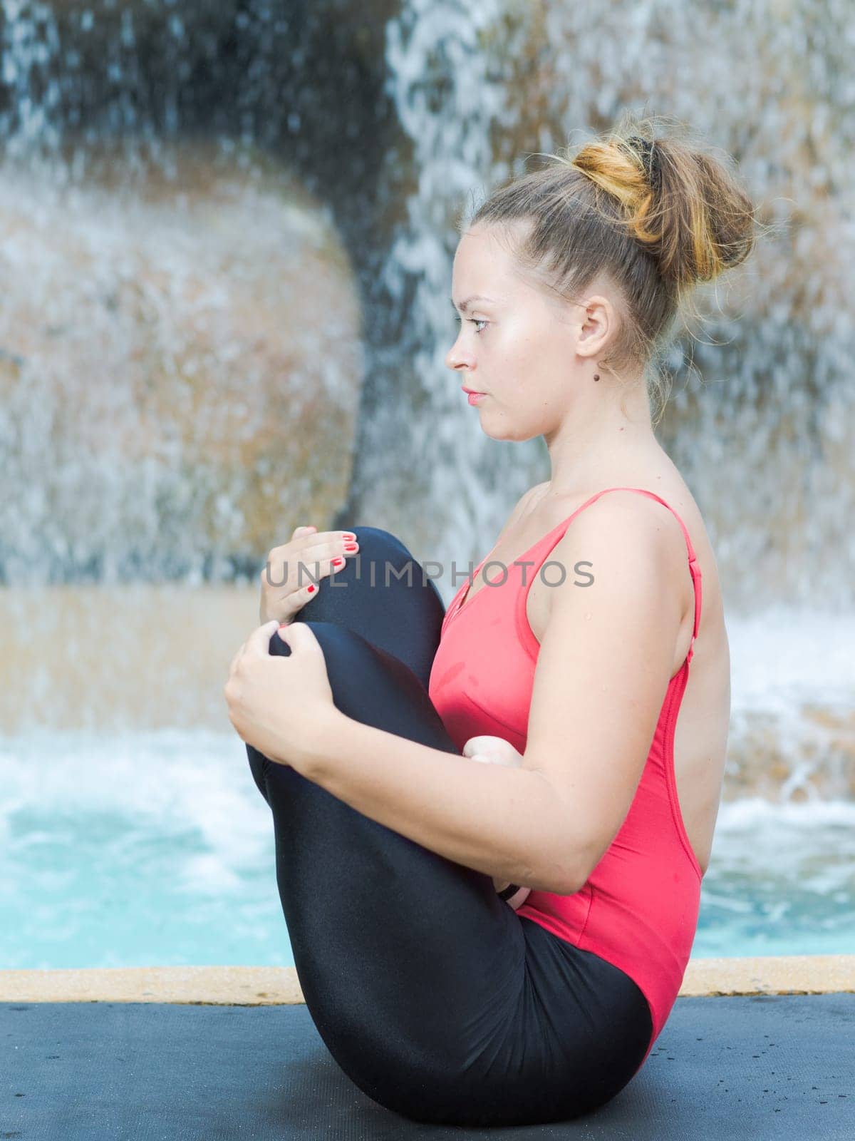 Woman practicing yoga near waterfall. Fetus Pose. Garbhasana