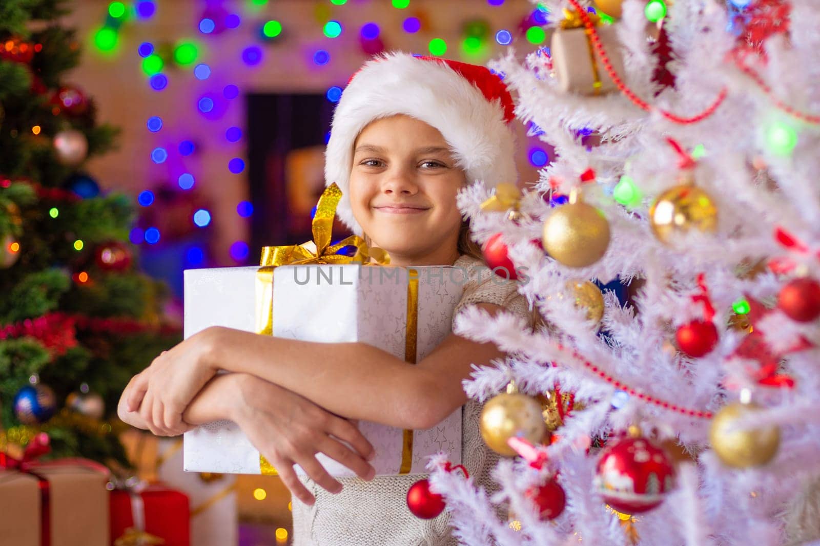 The girl at the Christmas tree holds a big gift in her hands