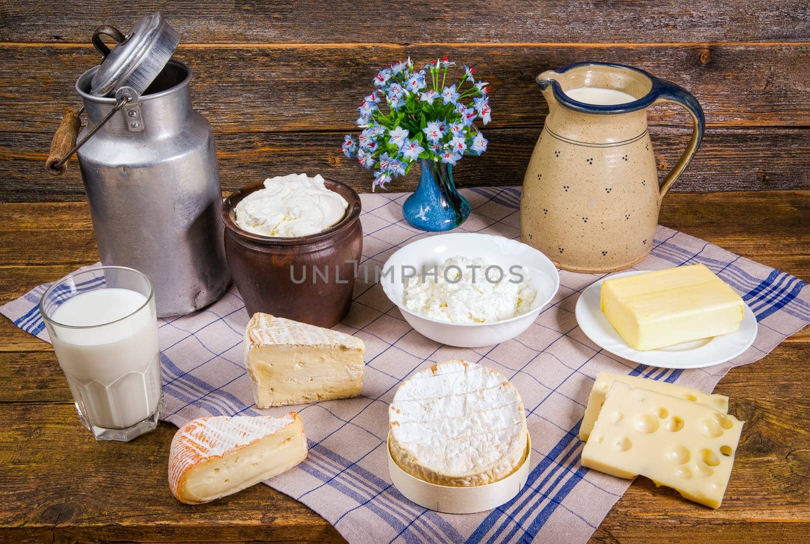 Milk in a jug and a glass, different sorts of cheese, whipped cream, curd, and a piece of butter in a rustic scenery on a wooden table with a kitchen towel.