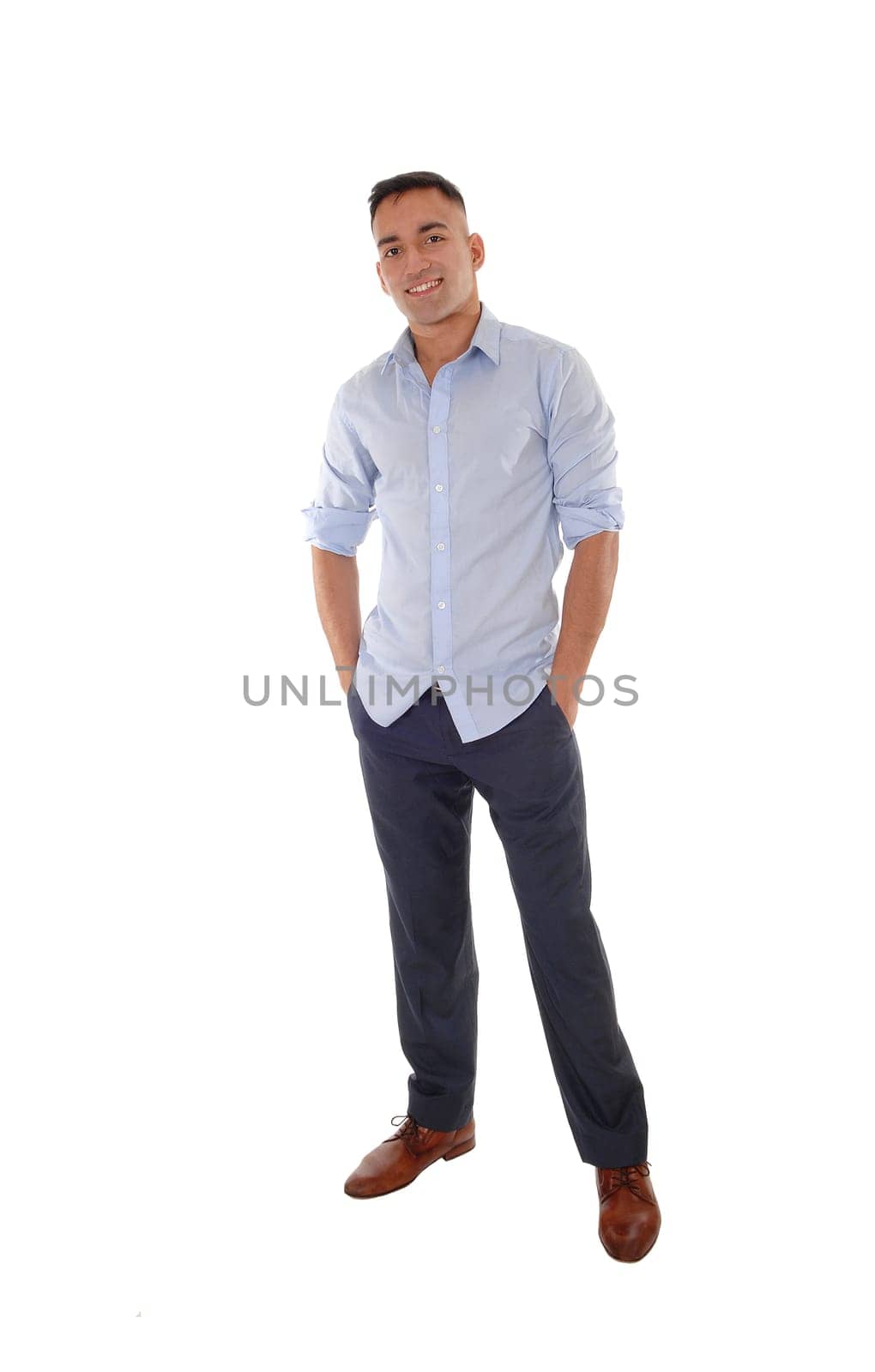 A handsome young east Indian man standing in a blue shirt with his.hands in his pocket, smiling, isolated for white background.