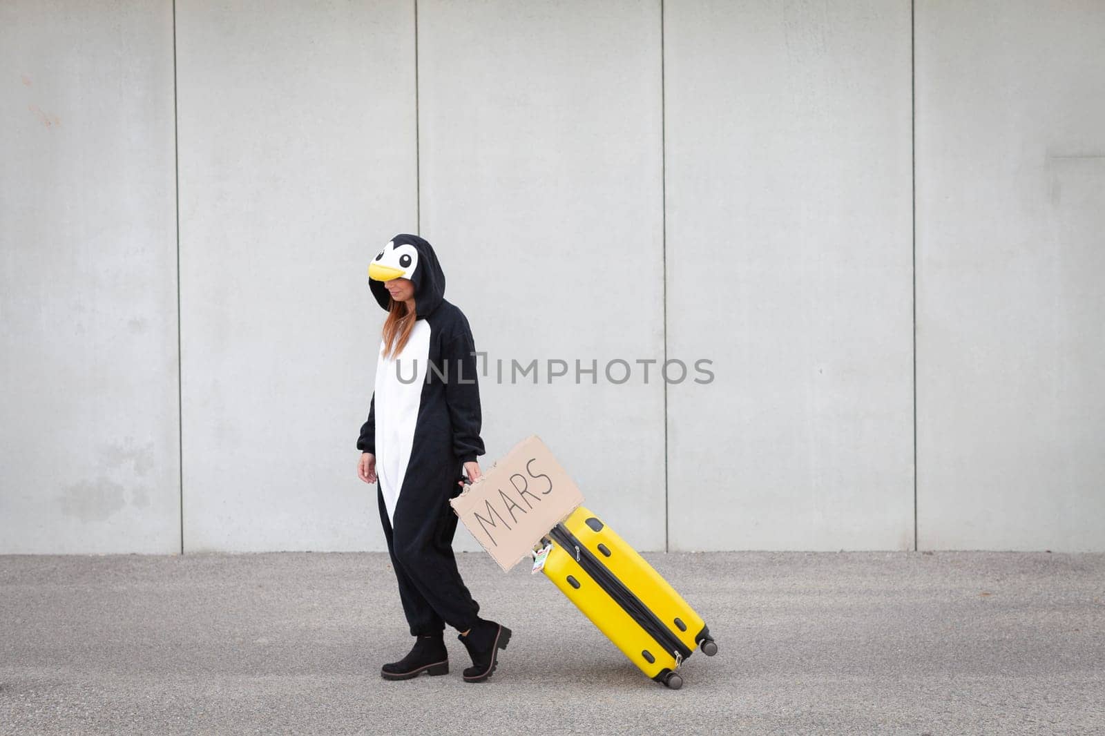 Young woman, with penguin costume and yellow suitcase, has to travel due to global warming. Symbolizes a sad penguin who has to leave his homeland because of global warming or climate change.